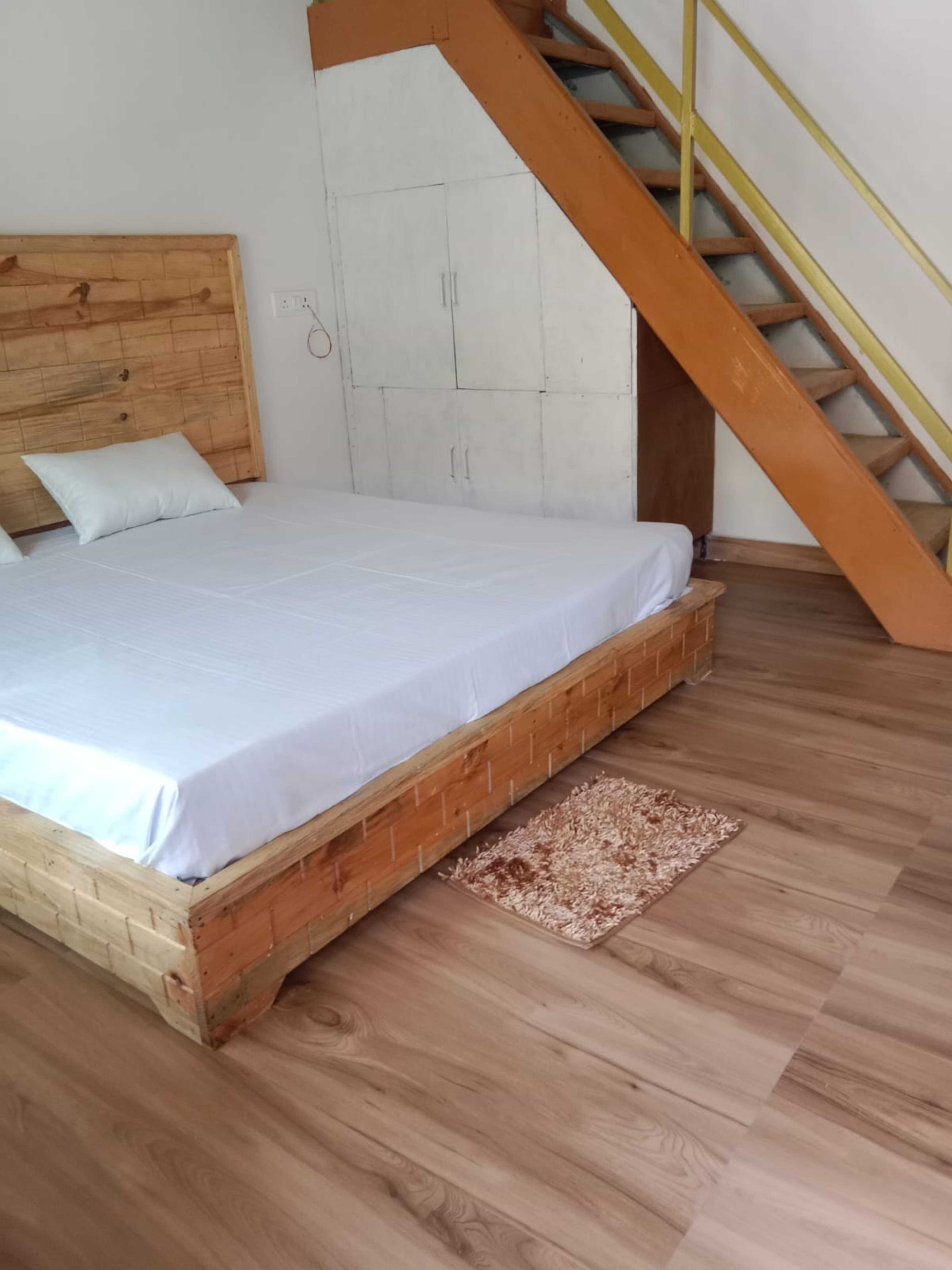 A bedroom with a wooden bed frame, a white mattress, a wooden headboard, a white wardrobe, and a wooden staircase with yellow handrails.
