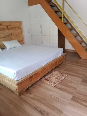 A bedroom with a wooden bed frame, a white mattress, a wooden headboard, a white wardrobe, and a wooden staircase with yellow handrails.
