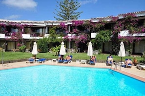 A swimming pool with lounge chairs and umbrellas, surrounded by a building with balconies adorned with flowering plants. - Casas Carmen (San Bartolomé de Tirajana)