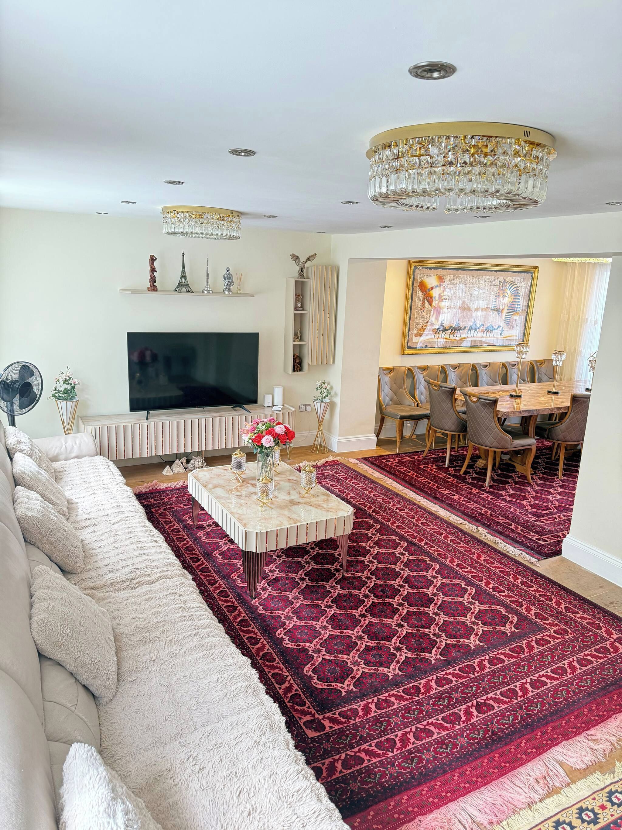 Un salon comprenant un canapé d’angle, un téléviseur à écran plat, un tapis rouge à motifs, un coin repas avec une table et des chaises, et un lustre.