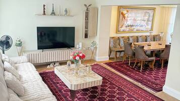 Un salon comprenant un canapé d’angle, un téléviseur à écran plat, un tapis rouge à motifs, un coin repas avec une table et des chaises, et un lustre.