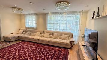 Een woonkamer met een hoekbank, een groot rood tapijt met patroon, een flatscreen televisie en decoratieve objecten aan de muur.