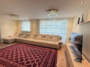 Een woonkamer met een hoekbank, een groot rood tapijt met patroon, een flatscreen televisie en decoratieve objecten aan de muur.