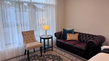 Una habitación de hotel con un ventanal grande, un sillón capitoné, una mesita redonda con una lámpara, un sofá rojo oscuro con almohadones decorativos y una alfombra estampada.