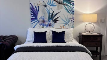Ein Schlafzimmer mit einem Bett, Kissen, einem Nachttisch, einer Lampe und einer Wand mit einem tropischen Gemälde.
