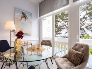 Ein Zimmer mit einem Glastisch, Stühlen, einer Lampe, einem Gemälde und Blick auf Bäume durch große Fenster.