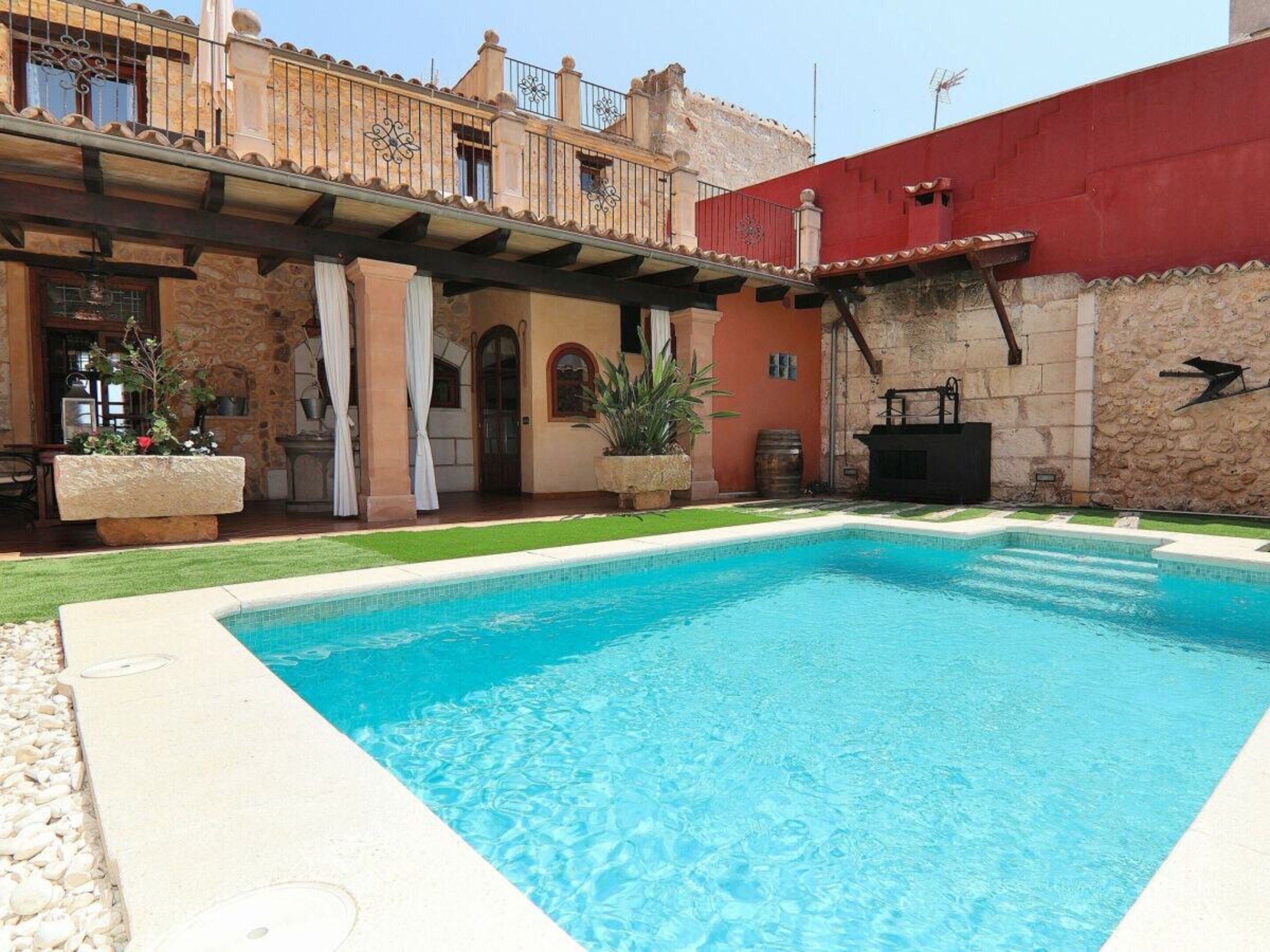 A swimming pool with a stone patio, a covered outdoor seating area, and a building with a red wall and stone facade.