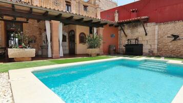 A swimming pool with a stone patio, a covered outdoor seating area, and a building with a red wall and stone facade.