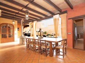 A spacious dining area with a long wooden table and chairs, a hanging light fixture, and a view of a stone wall and pool. - Villa in Muro With Swimming Pool (Muro)