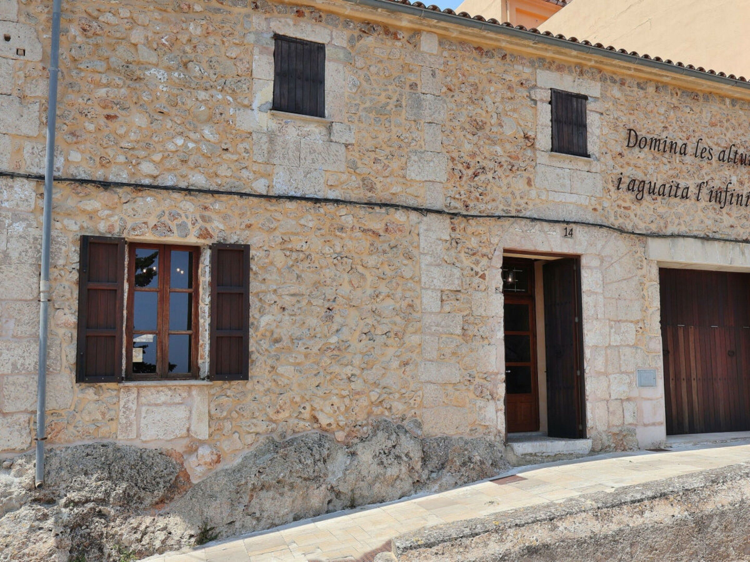 A stone building with a sign that reads 'Domina les altures laaguidat infin'. There are two windows with shutters and a door.