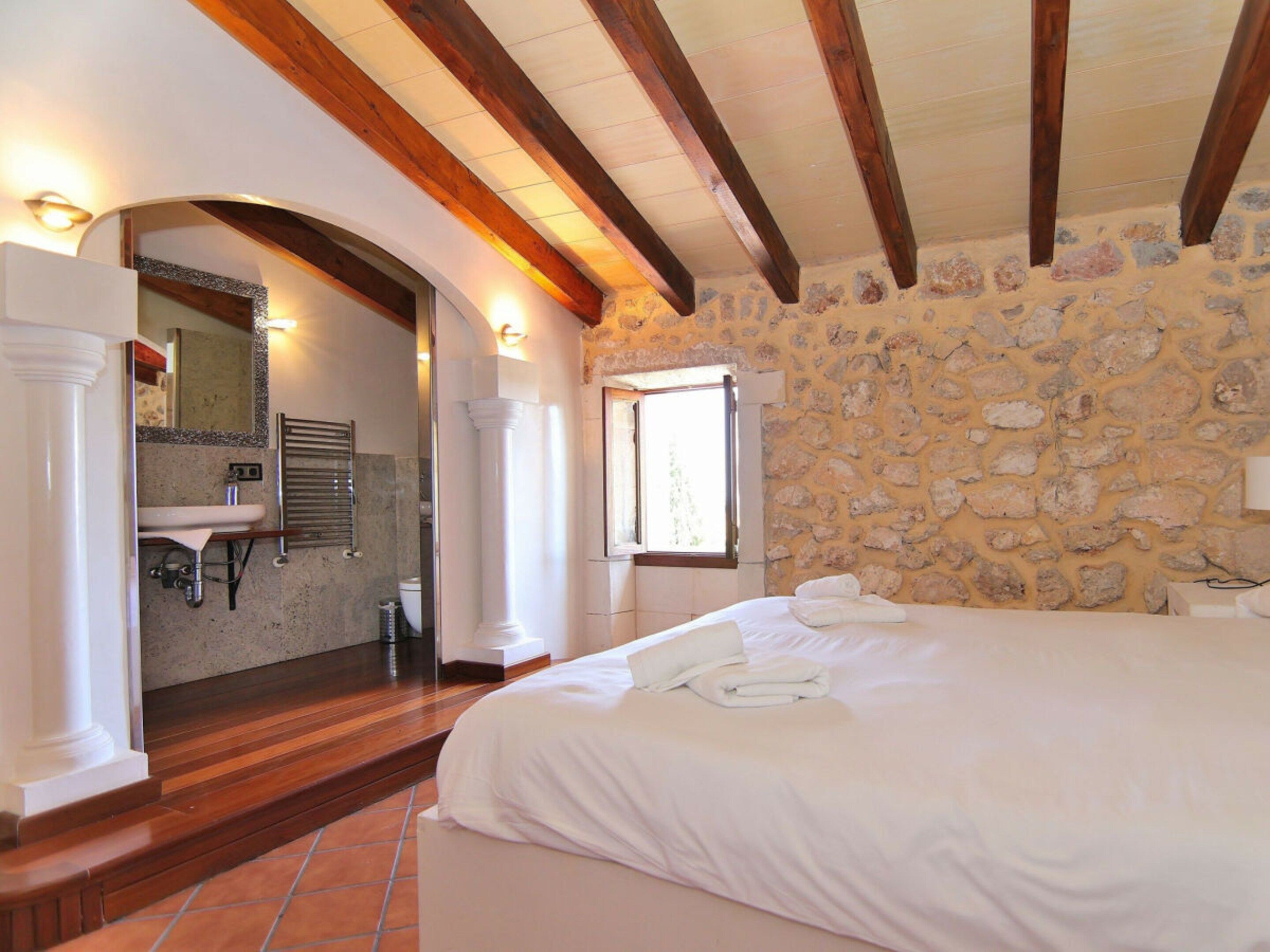 A bedroom with a stone wall, a wooden ceiling, a bed, a window, and a bathroom with a sink and mirror.