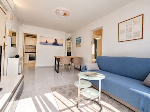 A modern living room with a blue sofa, a dining table with chairs, a kitchen in the background, and a ceiling light fixture. - Upper-floor bungalow with private solarium and community pool in San Luis (El Chaparral)