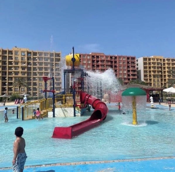 Parque acuático con un tobogán rojo y chorros de agua, rodeado de edificios de hotel.