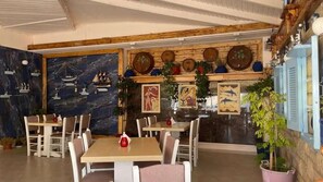 Interior de restaurante com mesas e cadeiras de madeira, um mural na parede e plantas em vasos.