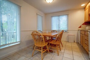 Uma área de refeições com mesa e cadeiras de madeira, uma cozinha com armários de madeira e janelas com persianas.
