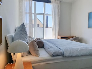 Quarto com cama, mesa de cabeceira, abajur e janela com cortinas.