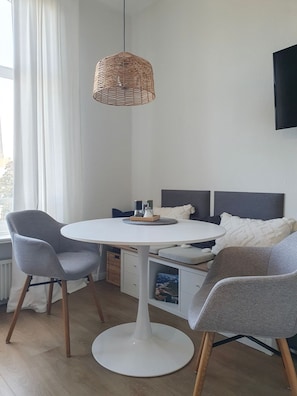 A modern room with a round white table, two grey chairs, a wicker hanging light fixture, a bed with grey headboard, and a television mounted on the wall. - "Mien lüttje Lichtblick" Vacation Rental—freshly renovated with a view of the (Borkum)
