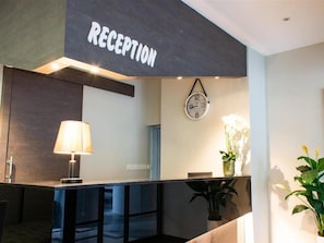 Un área de recepción con un cartel que dice 'RECEPTION', un reloj, una lámpara y plantas en macetas.
