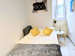 A bedroom with a bed, two pillows, a nightstand, a small table, and a window. - Sibenik Rituals Studio (Sibenik)