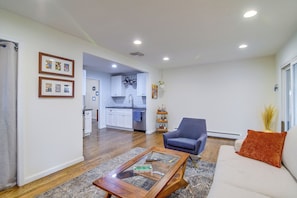Una sala de estar con un sofá, una mesa de centro y una silla. Se observa una cocina al fondo, con armarios blancos y un colgador para ollas.