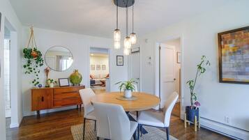 Un comedor con una mesa de madera, sillas blancas, luces colgantes y una planta en maceta.