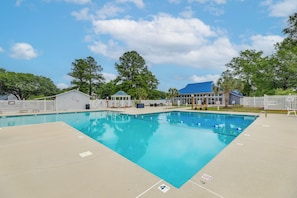 A rectangular swimming pool with a blue water surface, surrounded by a white fence, and a building with a blue roof in the background. - White Heron Landing - Waterfront New Bern Home! (New Bern)
