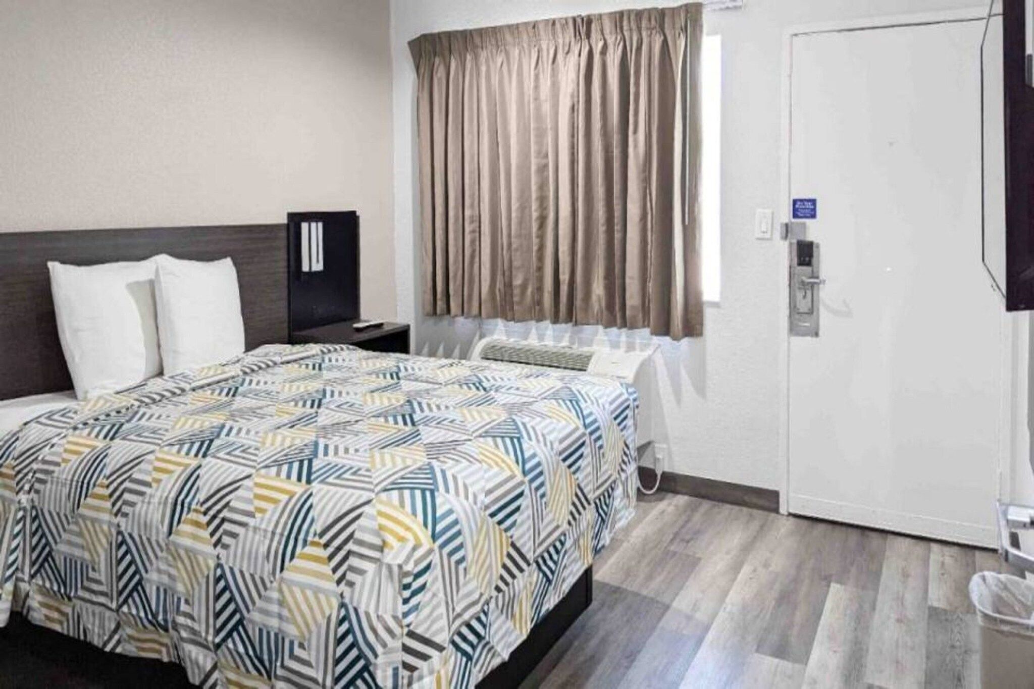 A hotel room with a bed, a nightstand, a window with curtains, a door, and a wall-mounted TV.