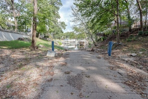 Un chemin menant à une zone délimitée par une barrière, avec une étendue d’eau, entourée d’arbres et d’un quartier résidentiel.