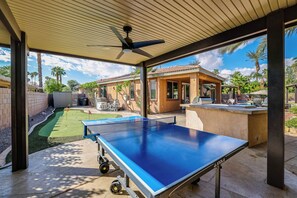 Una mesa de ping pong al aire libre con un ventilador de techo encima, rodeada de un patio con vista a una casa y palmeras.