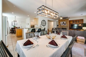 Un comedor moderno con una mesa grande dispuesta para cuatro, una cocina al fondo y una sala de estar con televisor y chimenea.