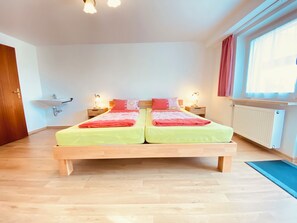 Een tweepersoonsbed met houten frame en een groene dekbedovertrek, een nachtkastje met een lamp, een wastafel aan de muur en een raam met gordijnen.