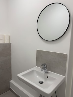 A bathroom with a white sink, a round mirror, and a toilet paper holder. - New & cozy in the heart of the city (Hamburg)