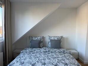 A bedroom with a sloped ceiling, a bed with a floral-patterned bedspread, two bedside tables, and a window with curtains. - New & cozy in the heart of the city (Hamburg)