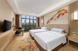 A hotel room with two beds, a TV, a desk, and a large window with a city view.