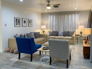 A living room with two sofas, a coffee table, and a blue chair. There are two lamps on side tables, a ceiling fan, and a window with curtains. - The O Spa + Resort, Adults Only (Desert Hot Springs)