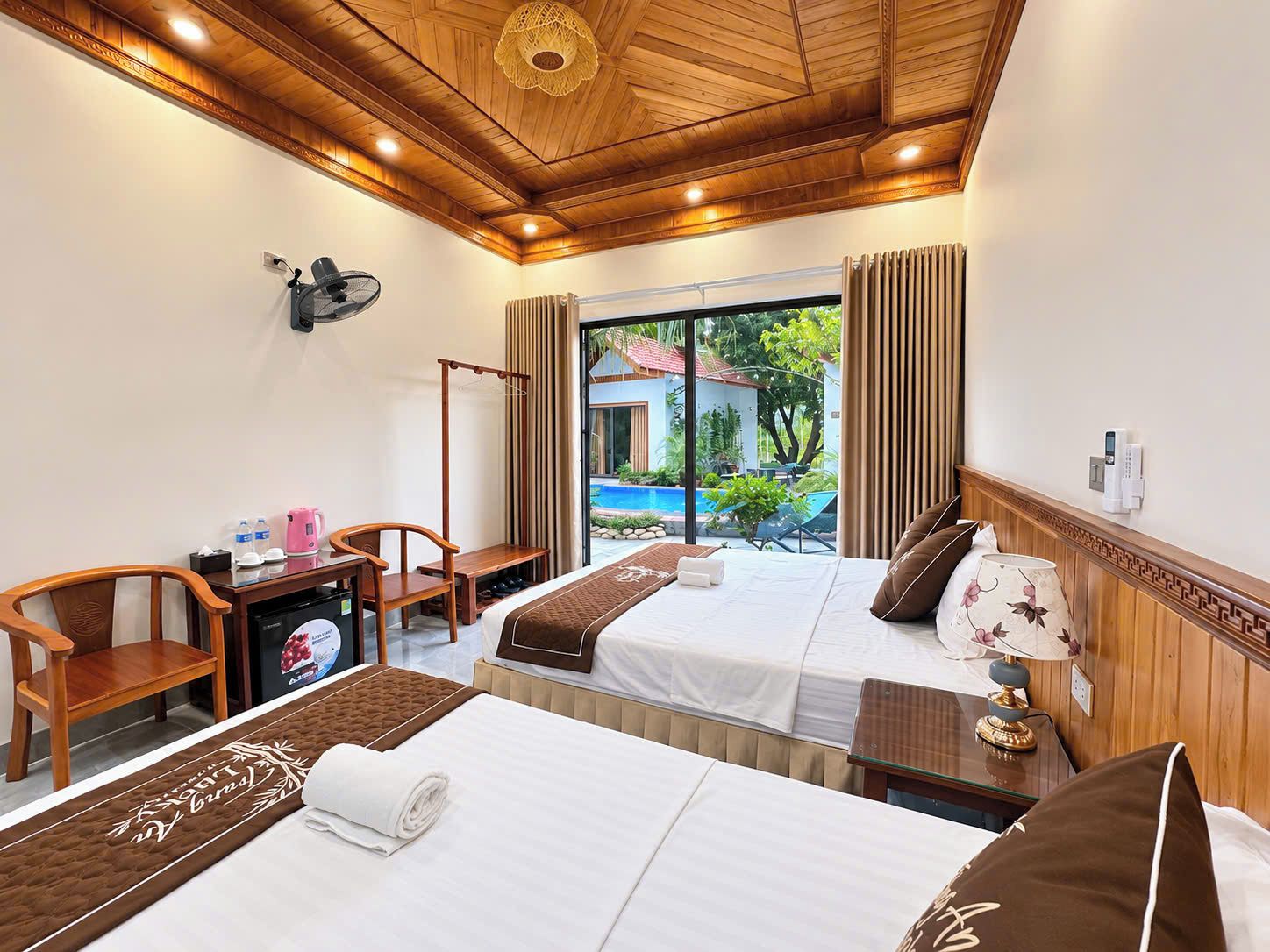 A hotel room with two beds, a desk, a chair, a fan, and a view of a swimming pool.