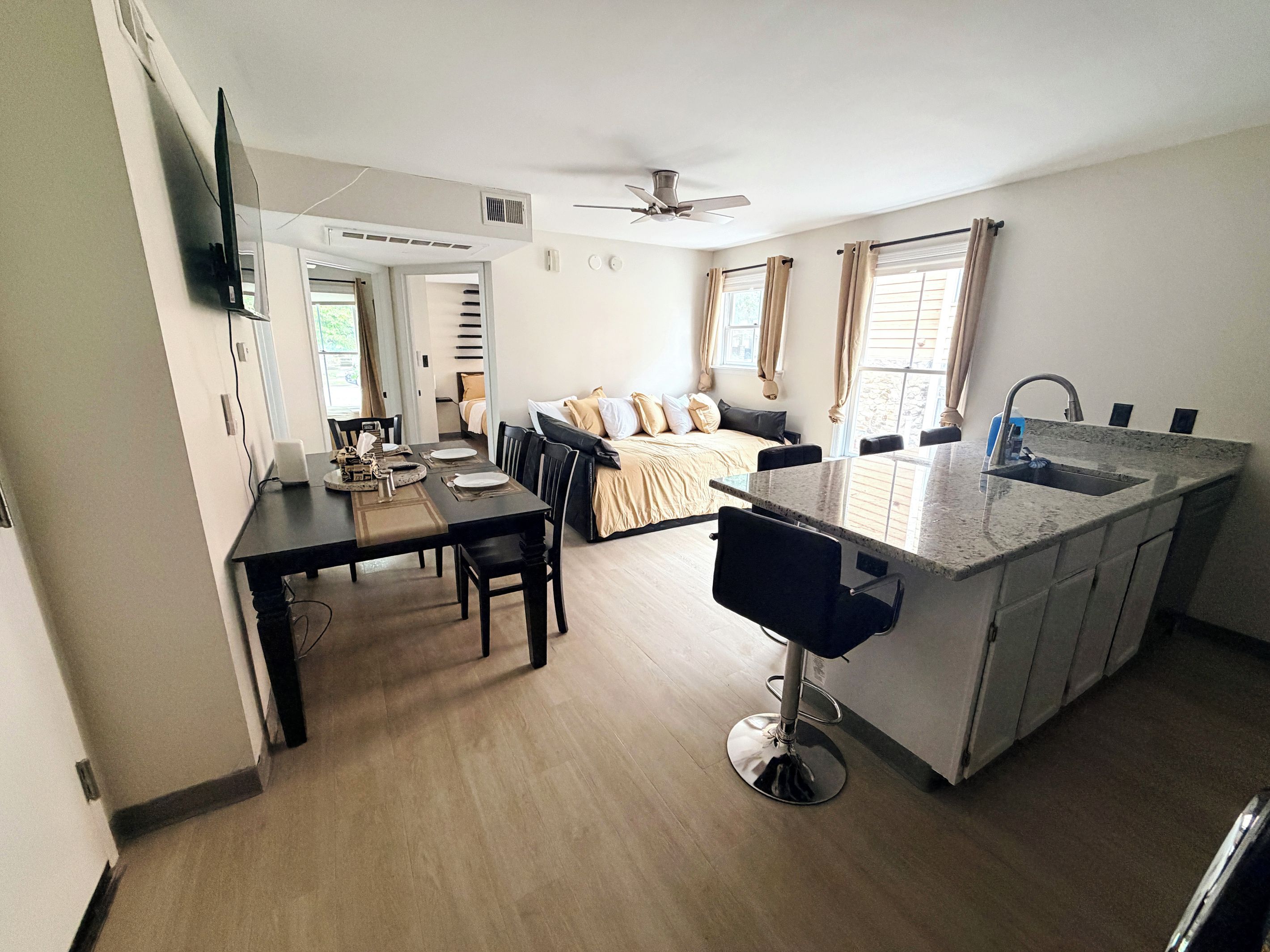 A modern kitchen with a central island, dining area, and a bedroom with a bed and curtains.