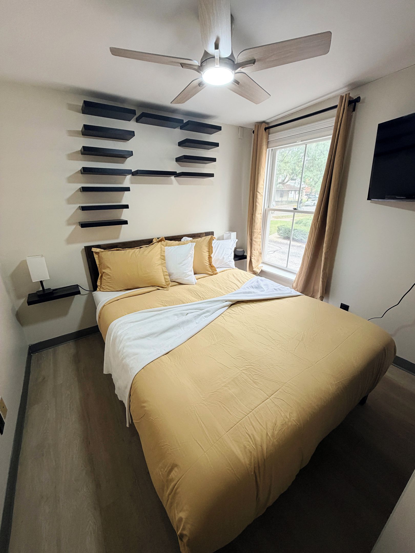 A hotel room with a bed, a ceiling fan, a window with curtains, and a wall-mounted TV.
