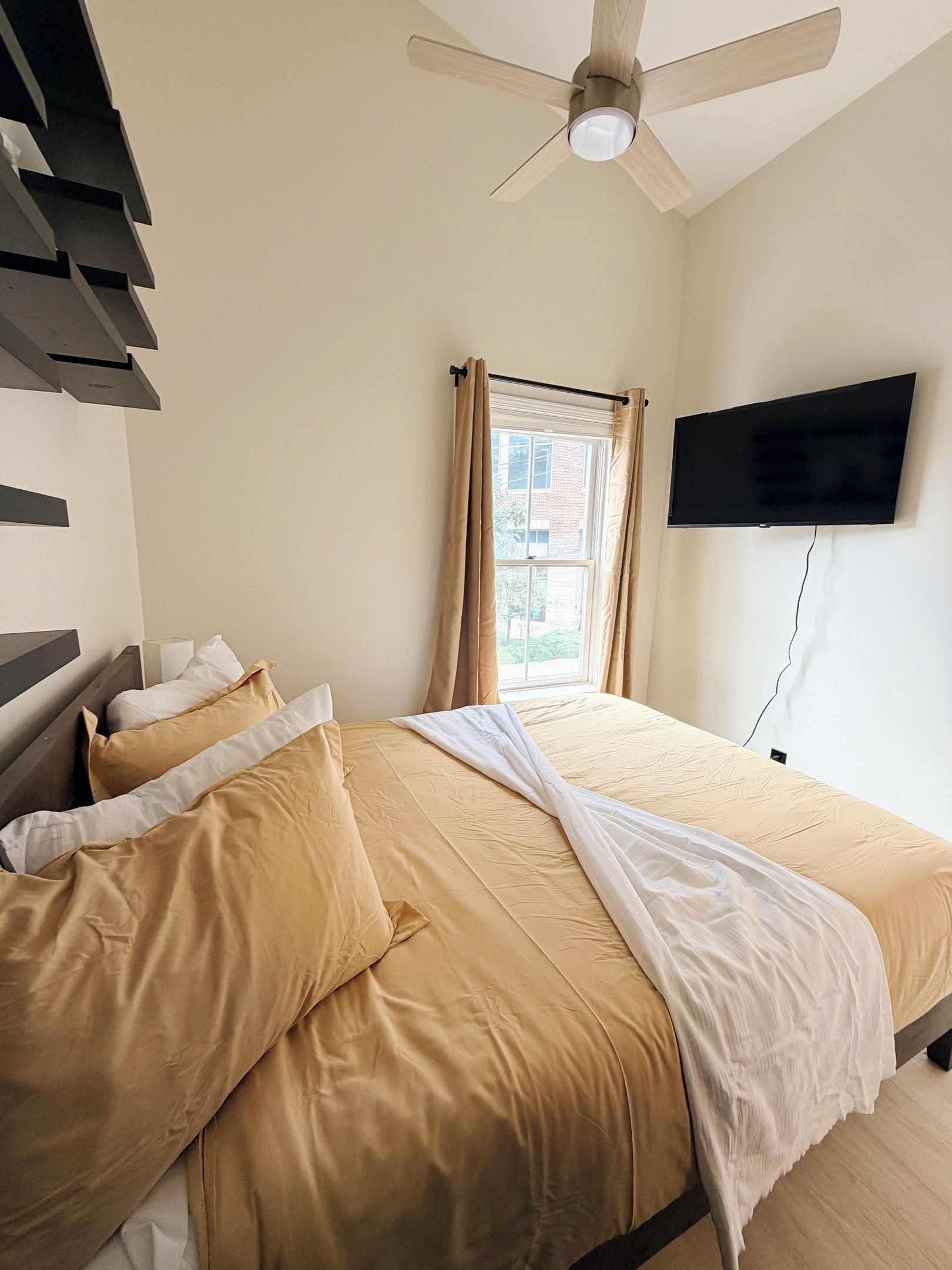 A bedroom with a bed, a ceiling fan, a window with curtains, and a mounted television.
