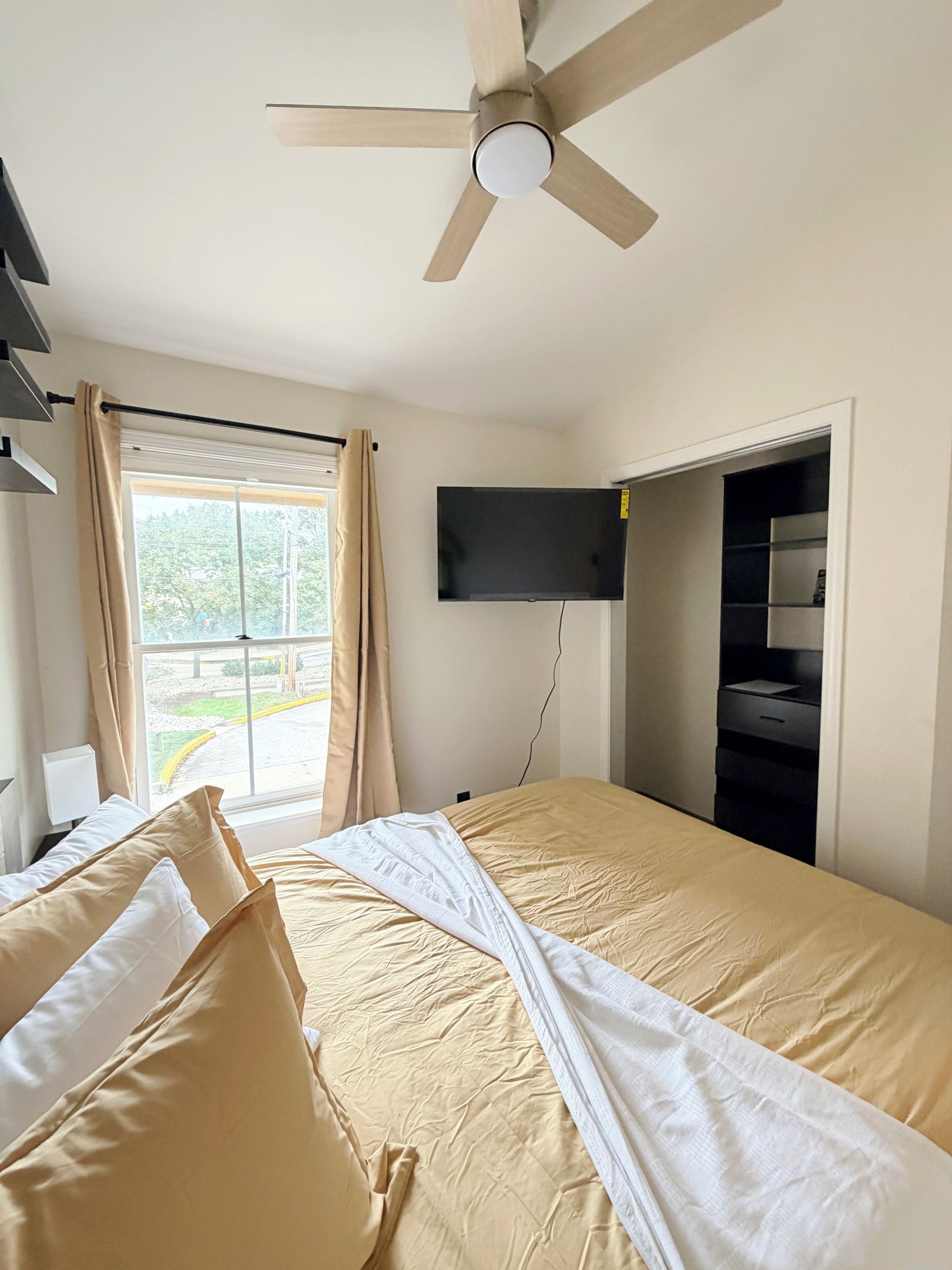 A hotel room with a bed, a ceiling fan, a TV mounted on the wall, and a window with curtains.