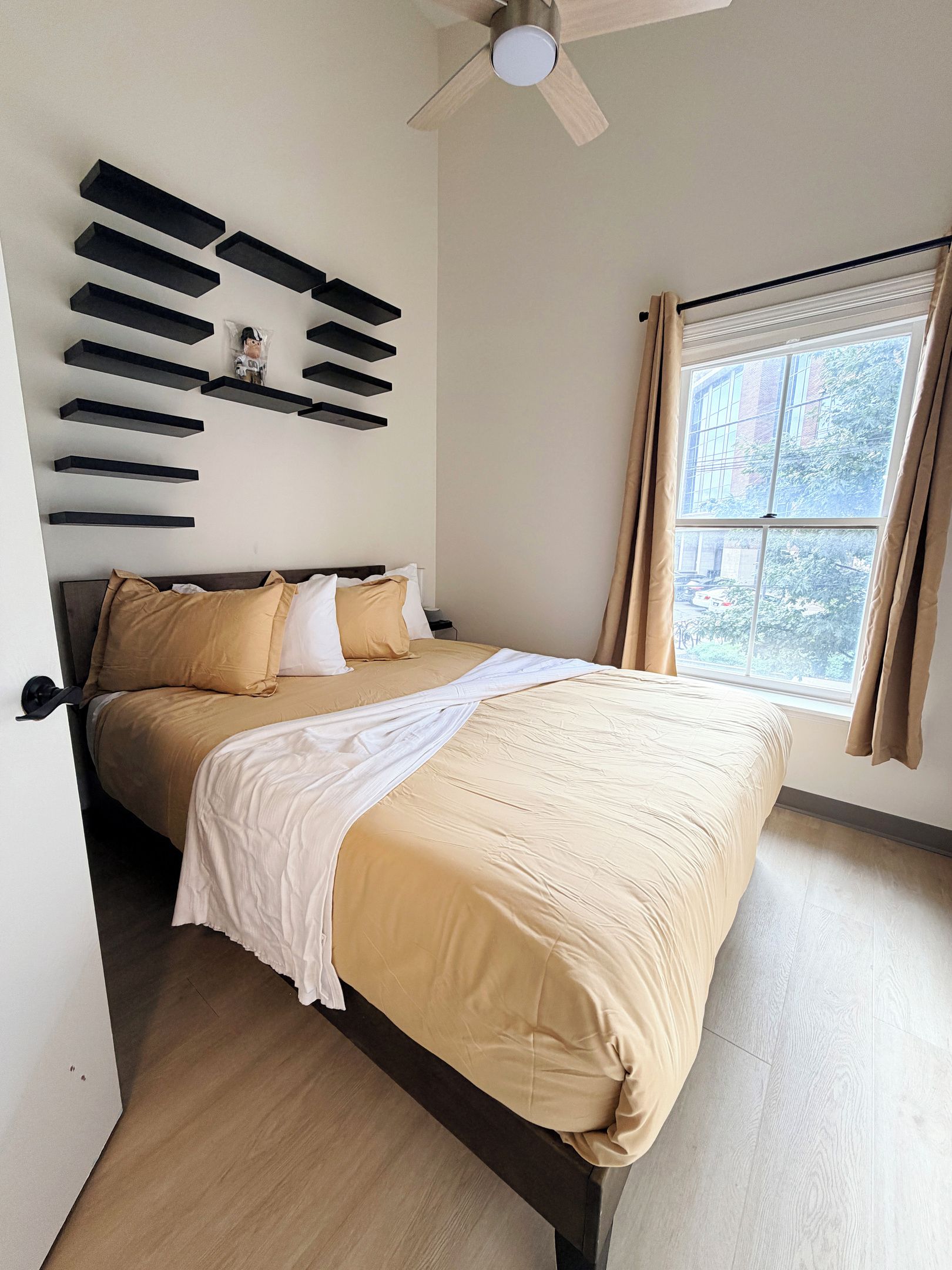 Un dormitorio con cama, ventilador de techo, ventana con cortinas y estante de pared con una figurita.