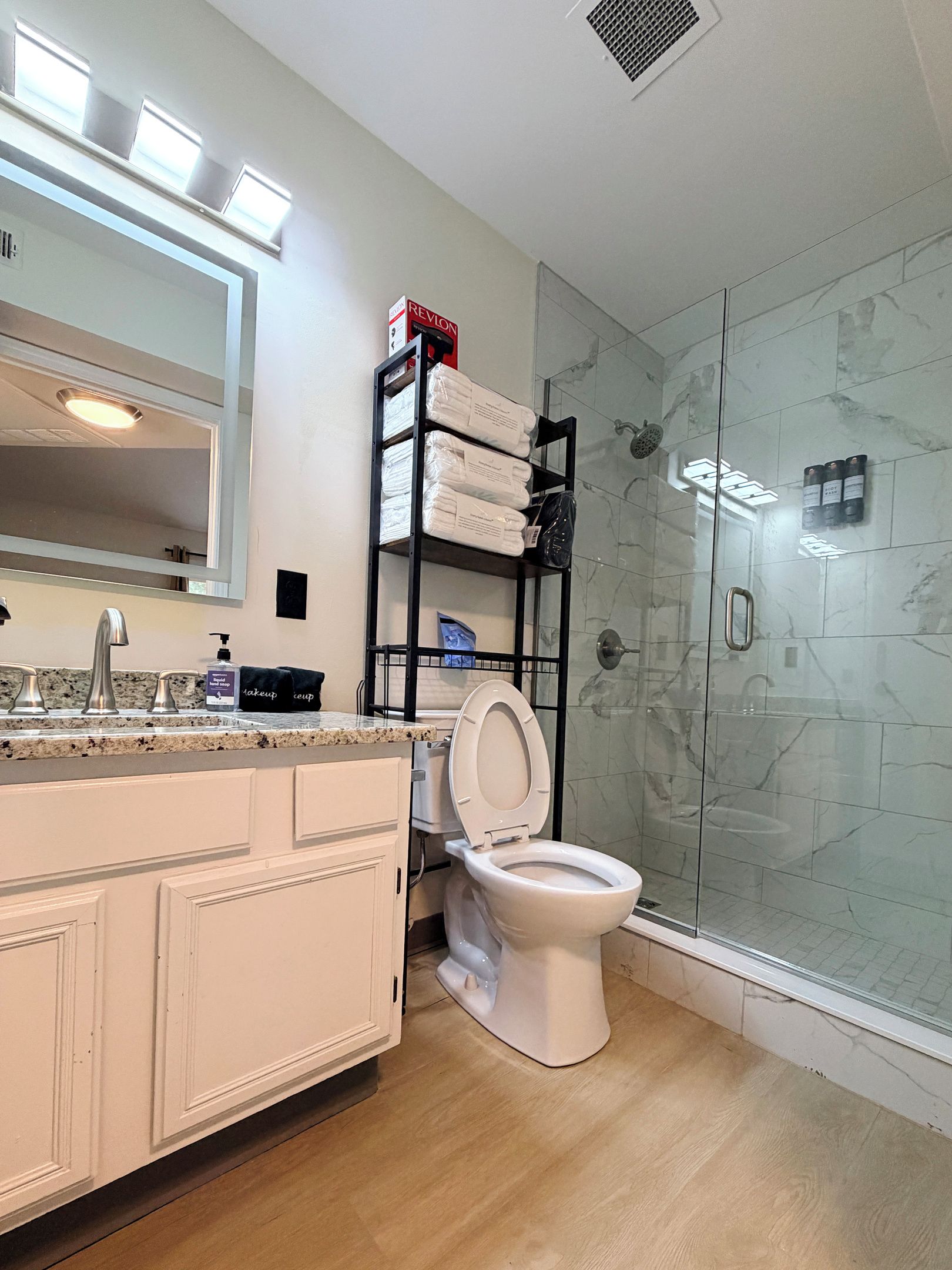 Un baño con cabina de ducha de vidrio, inodoro blanco, tocador con espejo y estante con artículos de aseo.