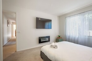 Un dormitorio con cama, un televisor de pantalla plana montado en la pared, una mesita con una planta y una ventana con cortinas translúcidas.