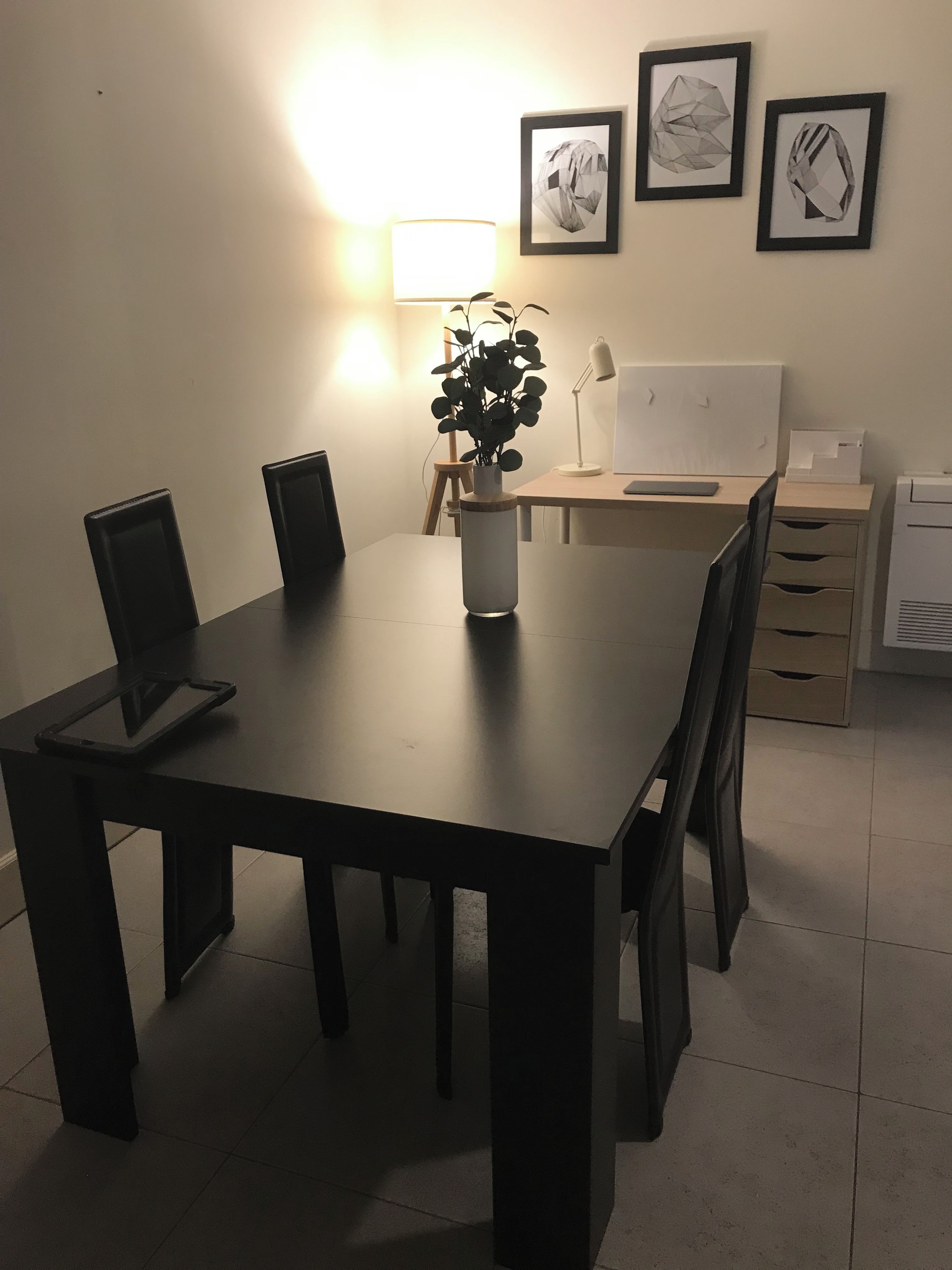A dining table with chairs, a plant, and a lamp. There are framed pictures on the wall.