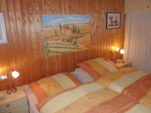 A room with wooden panel walls, a painting of a landscape, two bedside tables with lamps, and a bed with patterned bedding. - At the Broom Bush (Wettringen)