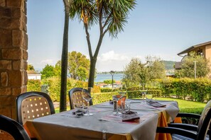 An outdoor dining area with tables set for two, overlooking a body of water and hills. - Apartment in Manerba With Barbecue (Manerba del Garda)