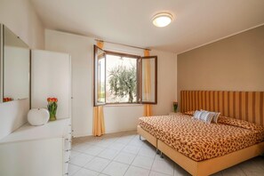 A bedroom with a bed, a dresser, a mirror, a window with curtains, and a view of trees outside. - Apartment in Manerba With Barbecue (Manerba del Garda)