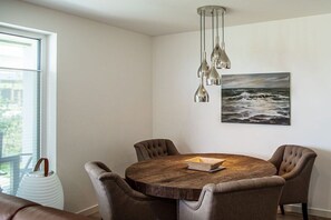 A dining area with a round wooden table, four upholstered chairs, a hanging pendant light, and a framed painting of a seascape. - Second Home Apartment 2 (Lohme)