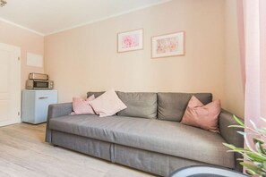 Ein Wohnzimmer mit einem grauen Sofa, zwei gerahmten Bildern an der Wand, einem kleinen blauen Schrank mit einer Mikrowelle und einer Tür links.