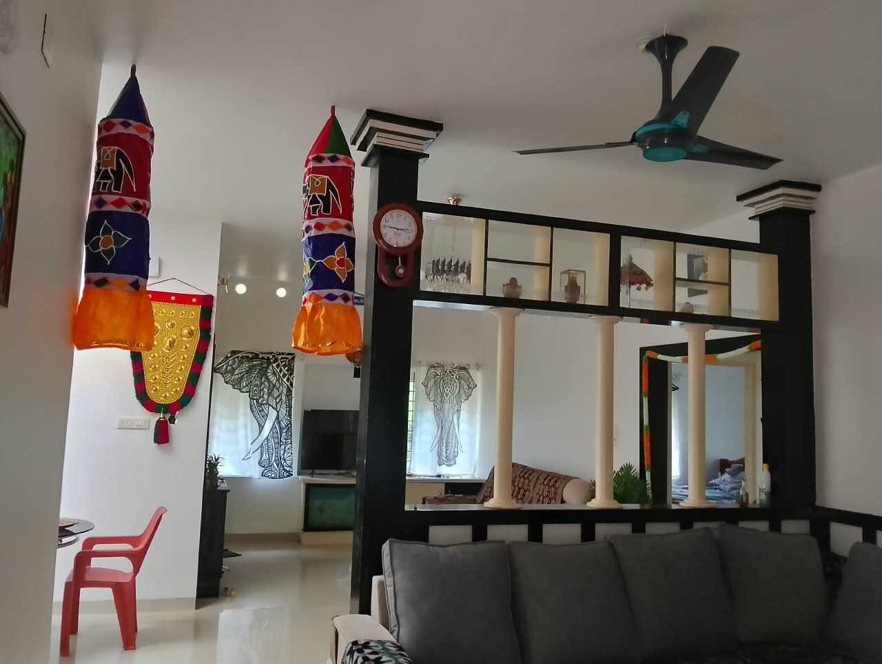 Una sala de estar con un sofá, una butaca roja, un ventilador de techo y objetos decorativos como un reloj y un espejo.